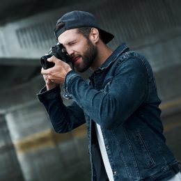 Photography hobby. Young stylish man standing on city street taking photos on camera concentrated side view